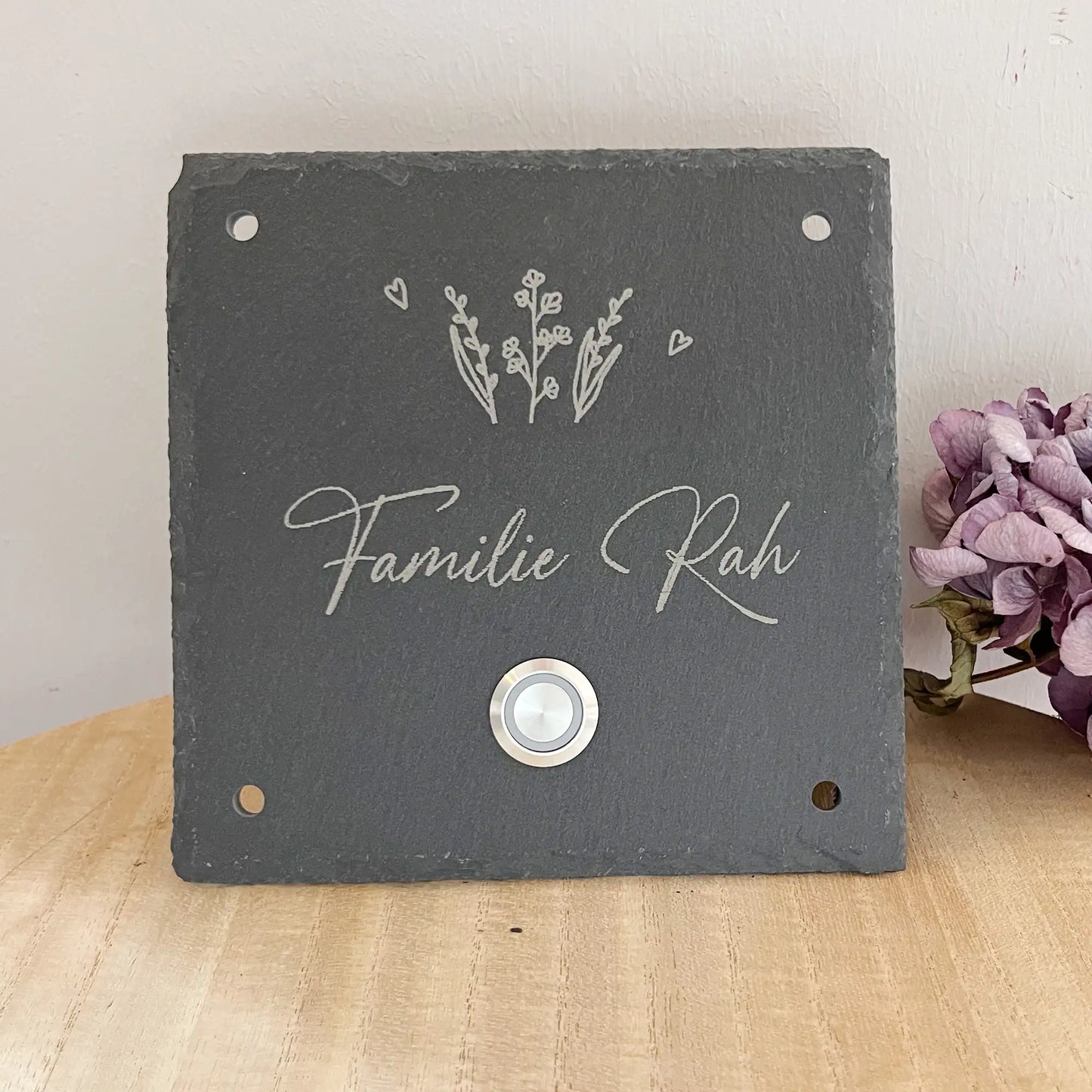 Personalisiertes Klingelschild aus Schiefer mit Edelstahl Taster mit oder ohne LED-Beleuchtung. Zarter Pflanzen oberhalb der kleinen Zeile für "Familie" oder "Willkommen bei" oder "Vornamen" oder "Straßenname mit Hausnummer". Klingelknopf unterhalb der Hauptzeile mit Familiennamen in geschwungener Handschrift. Hier in 15 x 15 cm mit LED-Edelstahltaster und 42 Loch Ecke für die Befestigung an die Hauswand als Beispiel