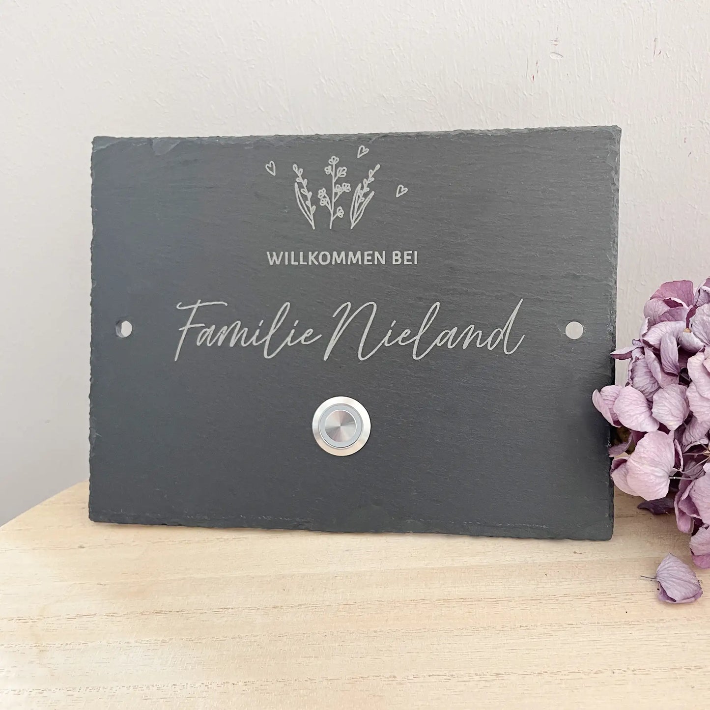 Personalisiertes Klingelschild aus Schiefer mit Edelstahl Taster mit oder ohne LED-Beleuchtung. Zarter Pflanzen oberhalb der kleinen Zeile für "Familie" oder "Willkommen bei" oder "Vornamen" oder "Straßenname mit Hausnummer". Klingelknopf unterhalb der Hauptzeile mit Familiennamen in geschwungener Handschrift. Hier in 20 x 15 cm mit LED-Edelstahltaster und 2 Loch Seite als Beispiel
