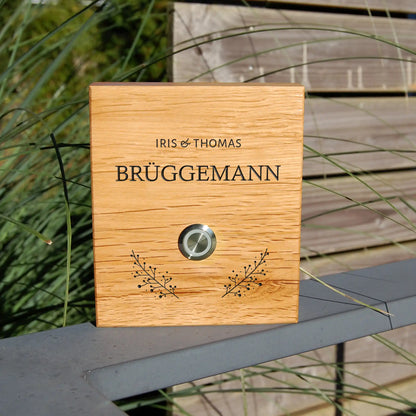 Klingelschild Motiv Brüggemann aus edler Eiche mit Tasterauswahl unbeleuchteter Taster, LED-Taster oder schwarz-Aluminium-Taster in der Größe 12 x 14 cm oder 14 x 25 cm. Name wird per Laser in das Holz graviert. Mit zwei grazilen Zweigen rechts und links unterhalb des Tasters und klassischer Schrift.