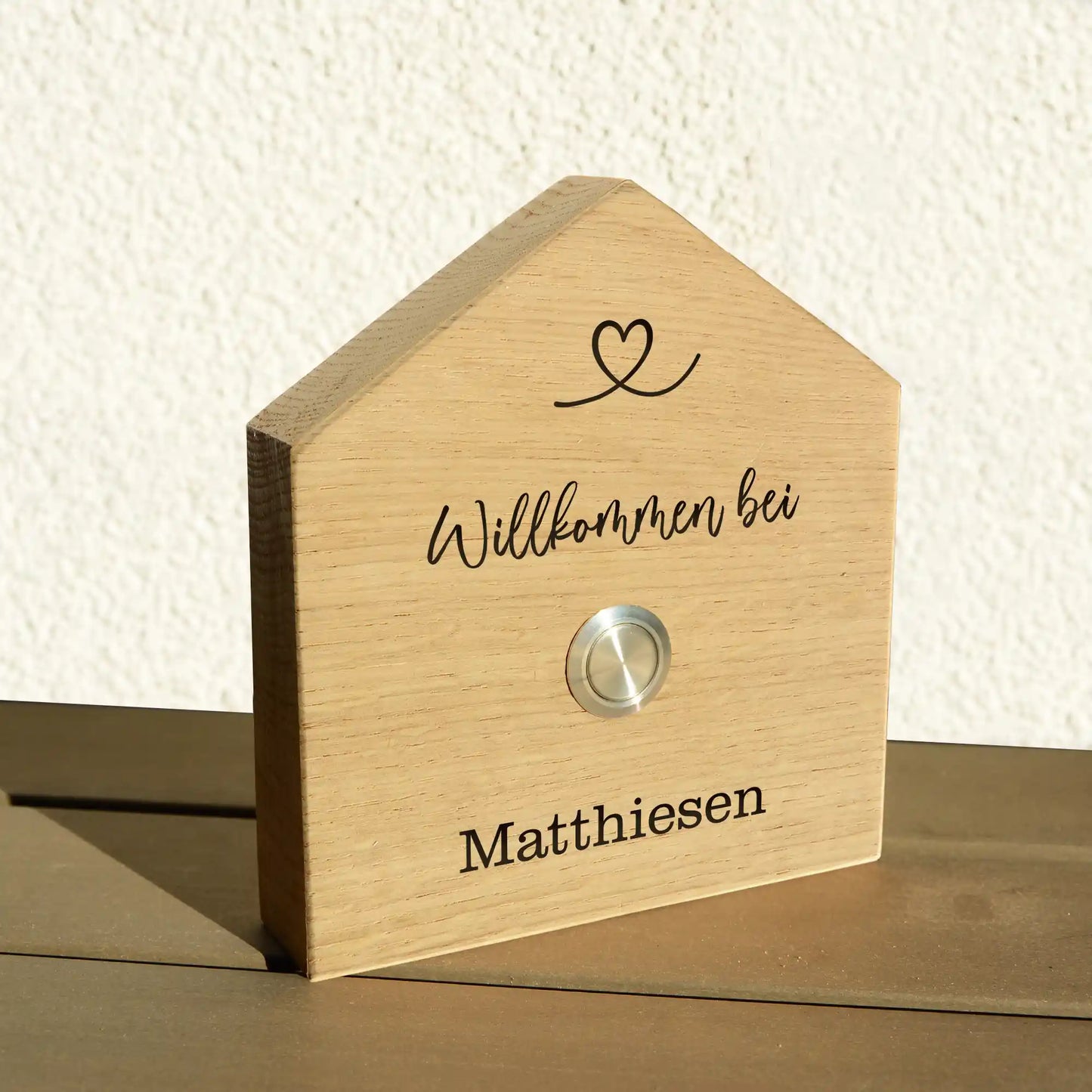 Klingelschild in Hausform aus edler Eiche mit Auswahl eines unbeleuchteten Tasters, LED-Taster oder schwarz-Aluminium-Taster in der Größe 12 x 14 cm oder 14 x 25 cm. Name wird per Laser in das Holz graviert. Mit einem geschwungenem Herz in der Dachspitze und in geschwungener Schrift "Willkommen bei" und unterhalb des Tasters "Familie" und der Nachname