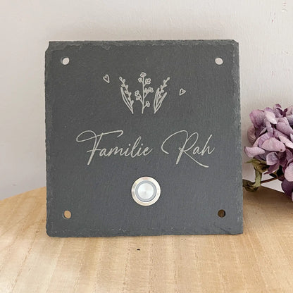 Personalisiertes Klingelschild aus Schiefer mit Edelstahl Taster mit oder ohne LED-Beleuchtung. Zarter Pflanzen oberhalb der kleinen Zeile für "Familie" oder "Willkommen bei" oder "Vornamen" oder "Straßenname mit Hausnummer". Klingelknopf unterhalb der Hauptzeile mit Familiennamen in geschwungener Handschrift. Hier in 15 x 15 cm mit LED-Edelstahltaster und 42 Loch Ecke für die Befestigung an die Hauswand als Beispiel