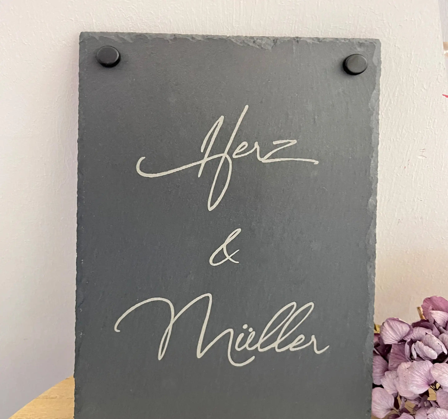 Tür- und Namensschild aus Schiefer mit schwungvoller Handschrift in personalisierter Form in der Größe 20 x 30 cm im Hochformat. Hier als Beispiel 2 Nachnamen mit "&"-Zeichen und schwarzen Abstandshaltern.
