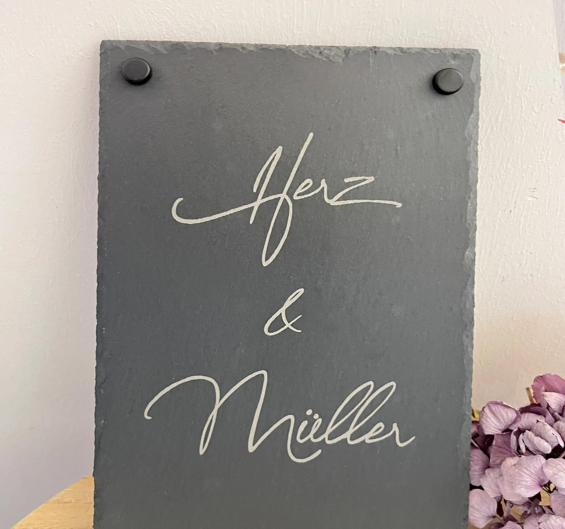 Tür- und Namensschild aus Schiefer mit schwungvoller Handschrift in personalisierter Form in der Größe 20 x 30 cm im Hochformat. Hier als Beispiel 2 Nachnamen mit "&"-Zeichen und schwarzen Abstandshaltern.
